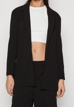 Mujer con un top corto blanco debajo de una chaqueta negra holgada y pantalones negros holgados, de pie frente a un fondo liso.