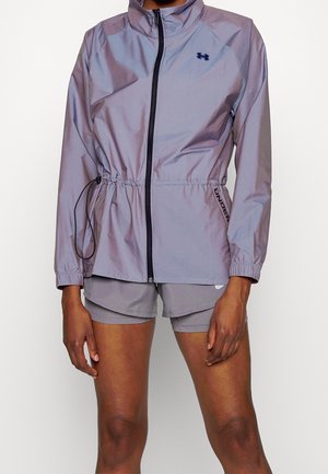 Person trägt eine hellviolette Zip-Up-Jacke mit elastischem Bund und passende Sportshorts, steht vor einem einfarbigen Hintergrund.