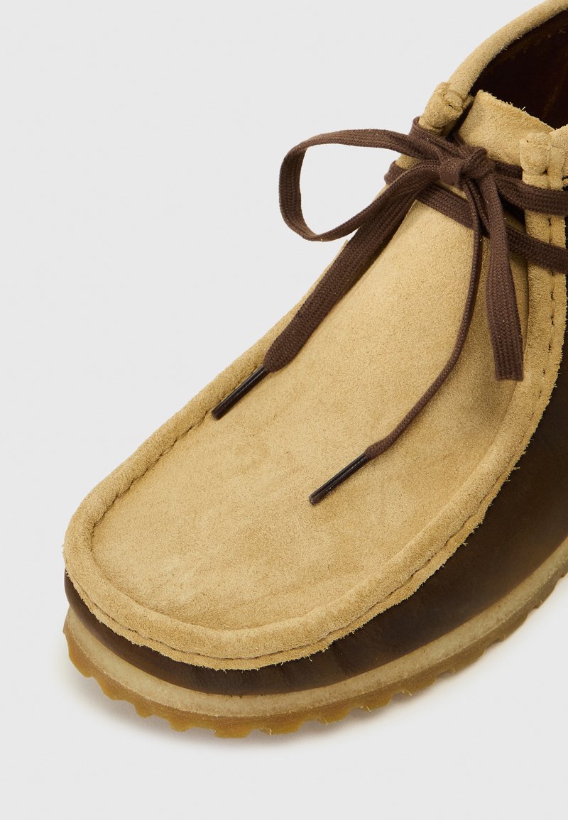 Chaussure en daim beige à bout arrondi, dotée de lacets marron et d'une semelle en caoutchouc. Tige texturée avec des détails de couture et un côté brillant.