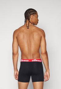 Svarta boxershorts med ett rött elastiskt midjeband som har "ellesse"-logotypen. Slät textur och åtsittande design, sedd bakifrån.