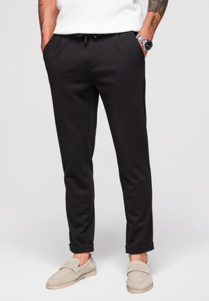 Mann trägt schwarze Slim-Fit-Hosen mit hochgekrempelten Bündchen, beige Loafers, weißes ärmelloses Oberteil, tätowierte Arme und eine Armbanduhr.