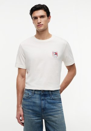 Νεαρός άνδρας που φοράει λευκό t-shirt Tommy Hilfiger με μικρό λογότυπο στο στήθος και μπλε τζιν, στέκεται μπροστά σε απλό λευκό φόντο.