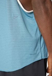 Camisola atlética azul da Nike, apresentando um tecido texturizado com pequenas perfurações, detalhes em branco e uma bainha curva. Logótipo "DRI-FIT" exibido.