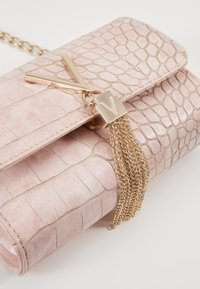 Růžová kabelka z imitace krokodýlí kůže se zlatým řetízkem, zdobená zlatým V-ozdobou a detailem s třásněmi.