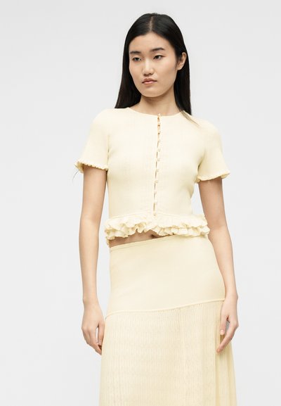 Νεαρή γυναίκα που φοράει κρεμ πουκάμισο με κοντά μανίκια, κουμπιά και βολάν στο τελείωμα, καθώς και φούστα με ψηλή μέση στον ίδιο χρωματισμό, στέκεται μπροστά σε μονόχρωμο φόντο.