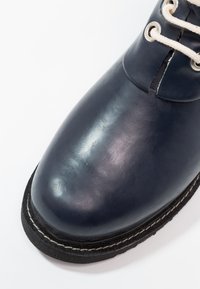 Bottine en cuir bleu marine avec une finition brillante, dotée de lacets blancs et d'œillets ronds en argent. Semelle en caoutchouc noire avec des détails de couture blanche.