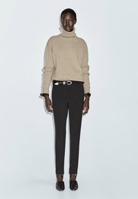 Maglione a collo alto beige, pantaloni neri aderenti con aperture alla caviglia e ballerine nere. Tessitura in maglia liscia, vestibilità rilassata, dettagli hardware minimi.