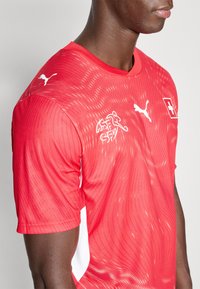 Piros sportpóló texturált mintával, fehér díszítésekkel és logókkal az elején és a vállán. Légáteresztő anyagból készült.