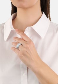 Anello d'argento con una fascia testurizzata, abbellito con piccoli pietre trasparenti, indossato su una mano vicino a una camicia bianca con colletto.