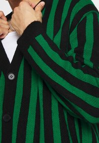 Detailní záběr na osobu, která si natahuje černozeleně pruhovaný cardigan s knoflíky přes bílou košili, přičemž je vidět stříbrný náramek.