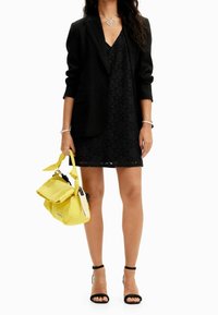 Robe en dentelle noire sous un blazer noir ajusté ; associée à des escarpins noirs à lanières et un sac à main jaune à plusieurs compartiments. Bijoux en argent en accent.