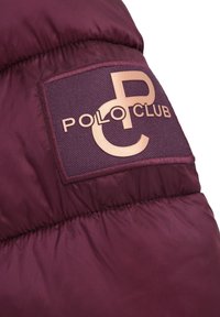 Vinrött quiltat tyg med en sydd applikation som visar stiliserade "PC"-bokstäver och texten "Polo Club" i metalliskt roséguld.