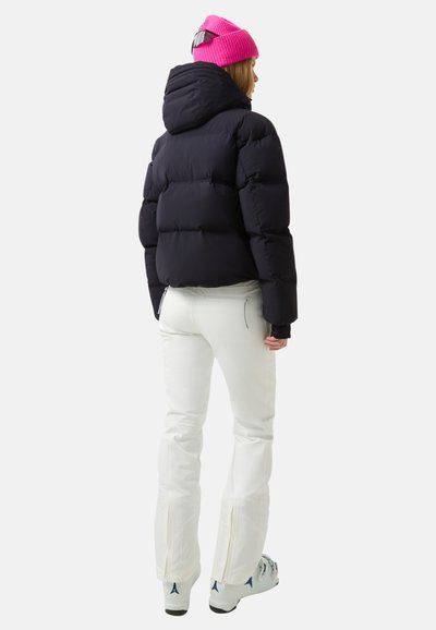 Zwarte gewatteerde jas met capuchon, gecombineerd met witte broek met ritsen bij de enkels. Een felroze gebreide muts maakt de look compleet.