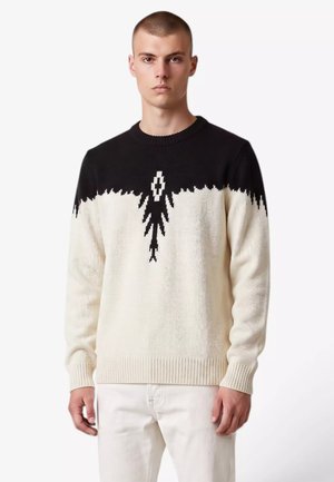 Giovane uomo che indossa un maglione bianco e nero con motivo geometrico e pantaloni bianchi, in piedi davanti a uno sfondo chiaro e uniforme.