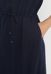 Robe bleu marine en tissu texturé avec une taille élastique et un cordon de serrage. Elle possède des poches latérales et des accents en métal argenté.