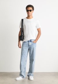 White cotton t-shirt with "Calvin Klein Jeans" logo, light blue denim jeans, black crossbody bag, sunglasses, and white sneakers.