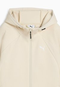 Beige Sportjacke mit Kapuze, ausgestattet mit einem durchgehenden Reißverschluss, strukturierten Seitenpaneelen und einem weißen Logo auf der linken Brust. Glattes Gewebe.