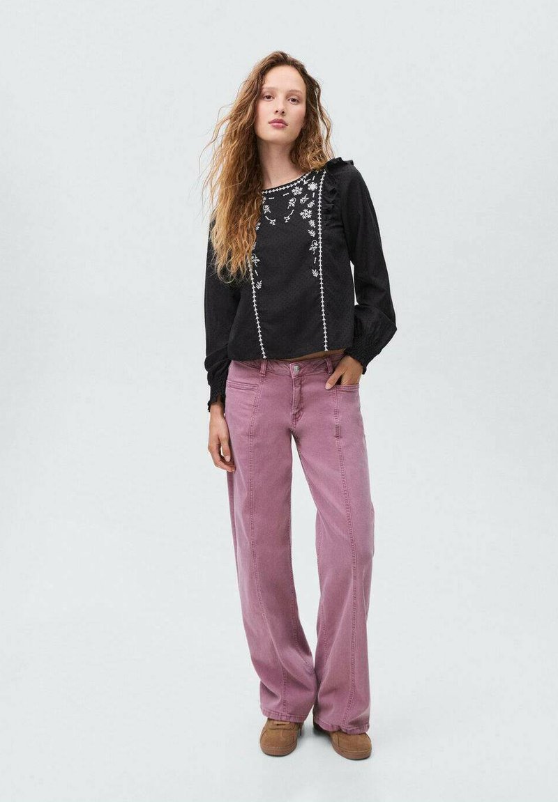 Blusa nera con texture e ricami floreali, maniche a volant, abbinata a pantaloni rosa chiaro a vestibilità ampia e calzature marroni.