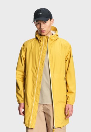 Hombre con un impermeable largo amarillo con capucha y una gorra negra, de pie contra un fondo gris liso.