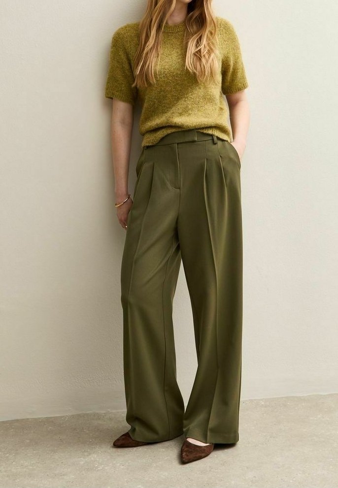 Femme debout portant un pantalon large vert olive, un pull court à manches courtes jaune moutarde duveteux, des chaussures pointues marron, devant un mur clair.
