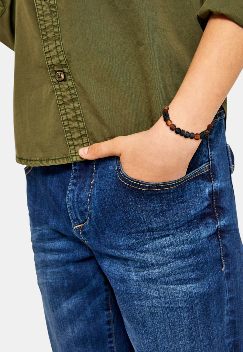 Camicia di cotone verde con bottoni, indossata sopra jeans in denim blu con cuciture gialle. Bracciale di perline marroni e nere al polso.