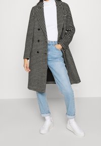 Abrigo de pata de gallo en blanco y negro con diseño de doble botonadura, grandes solapas y bolsillos laterales, combinado con jeans de color azul claro y zapatillas blancas.