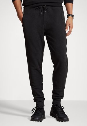 Homme portant un pantalon jogger noir avec cordon de serrage et poches latérales zippées, associé à des baskets noires et un haut noir à manches longues.