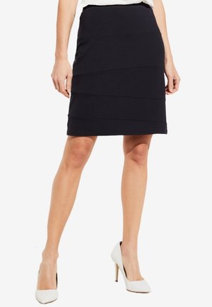 Femme portant une jupe noire longueur genou avec des coutures horizontales, associée à des chaussures blanches à talons hauts, debout les jambes légèrement écartées.