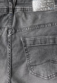 Vaqueros de denim gris con un bolsillo de parche, costuras detalladas y una etiqueta plateada texturizada en la cintura. Superficie de tela suave.