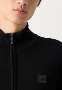Black zip-up sweater with a high collar, ribbed texture, and a square logo patch on the chest featuring embossed lettering.