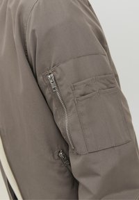 Manga de chaqueta marrón con dos bolsillos con cremallera y un bolsillo rectangular cosido sobre un fondo claro.