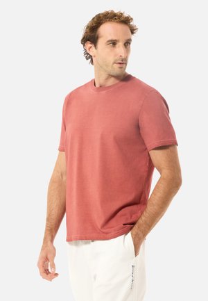 Homme portant un T-shirt rouge délavé à manches courtes et un pantalon blanc, une main dans la poche, regardant vers sa droite sur un fond uni.