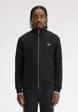 Man with braided hair wearing a black zip-up jacket with white logo on chest and striped cuffs, standing against a plain background.