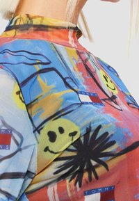 Colorful mesh top featuring a mix of blue, yellow, and red. Design includes smiley faces, abstract shapes, and brand tags.