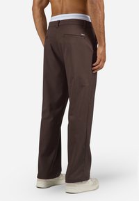 Muški model u tamnosmešnim ravnim hlačama i belim patikama, prikazan s leđa bez majice na beloj pozadini.