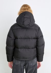 Svart dunjakke med hette, med quiltet design og ribbede mansjetter. Materialet har en glatt, matt finish.