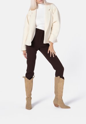 Kvinde poserende iført cremefarvet uldfrakke, hvid rullekrave, mørkebrune bukser og tanfarvede ruskind knæhøje støvler med lave hæle.