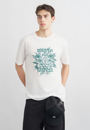 Νέος άνδρας που φοράει λευκό μπλουζάκι με πράσινα μάτια και το κείμενο "Earth Tripper", κρατώντας μαύρο καπέλο με δίχτυ, στέκεται μπροστά σε απλό γκρι φόντο.