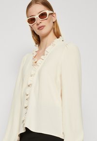 Long-sleeve cream blouse with ruffled trim along the v-neckline. Soft fabric and loose fit. Model wearing light-colored sunglasses.