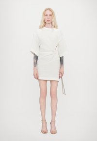 White short dress with short puff sleeves, featuring a wrap design, textured fabric, and a tied detail at the waist. Model wearing nude sandals.
