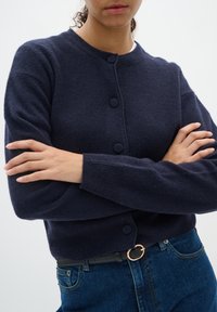 Mörkblå cardigan i ullblandning med rund hals, långa ärmar och fyra knappar fram. Kombineras med högmidjade blå jeans och ett bälte.