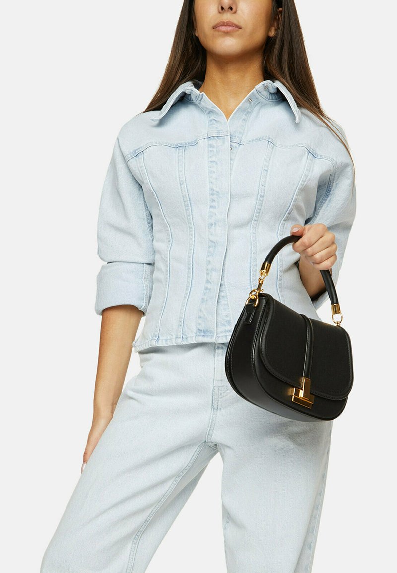 Borsa in pelle nera con forma strutturata, con accessori dorati e manico superiore, tenuta da una persona che indossa una giacca di jeans chiara e pantaloni.