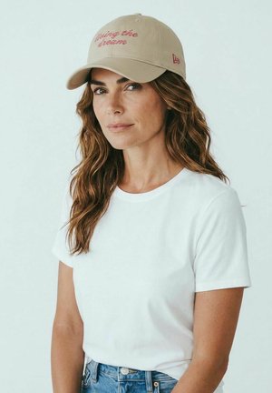 Woman with wavy brown hair wearing a beige baseball cap with "living the dream," a white t-shirt, and blue jeans, against a plain light background.
