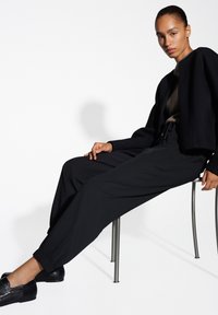 Giacca nera oversize con una parte superiore marrone testurizzata, pantaloni larghi neri e scarpe nere slip-on. Modella seduta su una sedia di metallo.