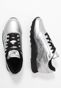 Stříbrné sportovní boty s hladkým kovovým povrchem, černými tkaničkami a gumovou podrážkou. Jsou opatřeny perforovanými detaily a logem na jazyku.