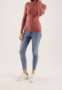 Gerippter Rollkragenpullover in staubigem Rosa, figurbetontes Design; kombiniert mit engen, hellblauen Jeans und weißen Sneakern mit goldfarbenen Akzenten.