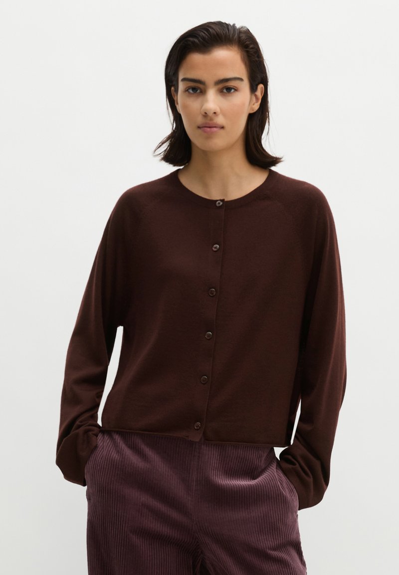Young woman with short dark hair wearing a dark brown buttoned cardigan and purple corduroy pants with hands in pockets.