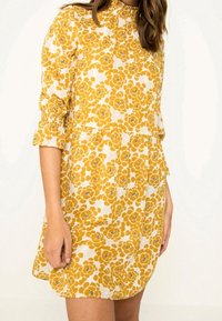 Vestido floral amarillo con fondo blanco, de mangas largas y puños fruncidos. La tela tiene un aspecto texturizado y un corte holgado.