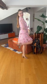 Γυναίκα με ροζ φορέμα με λουλούδια και μπεζ τακούνια ποζάρει με ένα πόδι σηκωμένο σε σαλόνι με καναπέ, τηλεόραση, μηχάνημα γυμναστικής και γλάστρα.
