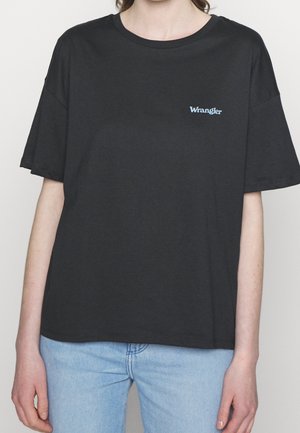 Personne portant un t-shirt Wrangler noir uni à manches courtes et un jean bleu clair, visible du cou aux hanches sur un fond blanc.